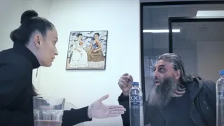 Twee mensen in gesprek aan een tafel in een kantoorruimte: een witte vrouw met strak naar achter gebonden donker haar en een oorbel gebaart naar een man met een donkere huidskleur, lange grijze baard en lang haar, die nadrukkelijk iets zegt. Op tafel staan glazen en flessen water. Aan de muur hangt een schilderij van twee vrouwen.