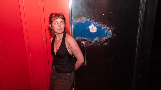 Een persoon met kort roodbruin haar, een froufrou en een zonnebril op het hoofd leunt tegen een zwarte deur in een felrood verlichte ruimte. De persoon kijkt opzij en draagt een zwarte top met een diepe V-hals, een zilveren ketting en een donkere broek. Op de linkerarm zijn fijne tatoeages te zien. De zwarte deur heeft een metalen handvat en een grote, opvallende slijtplek waar blauwe verf en het onderliggende materiaal doorheen komen.