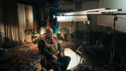 Een oudere man met een lichtbruine huid en grijs haar zit in een rijk gedecoreerd militair uniform voor een interview. Om hem heen staan lampen, microfoons en andere filmapparatuur; achter hem hangen vlaggen.