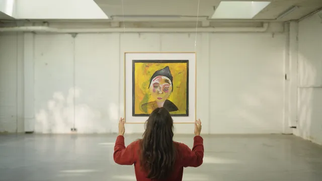 Een vrouw met lang zwart haar en een donkerrode trui, op de rug gezien, houdt een schilderij omhoog in een lege, lichte tentoonstellingsruimte. Het schilderij toont een abstract portret met gele achtergrond en een figuur met gesloten ogen en een zwarte muts.