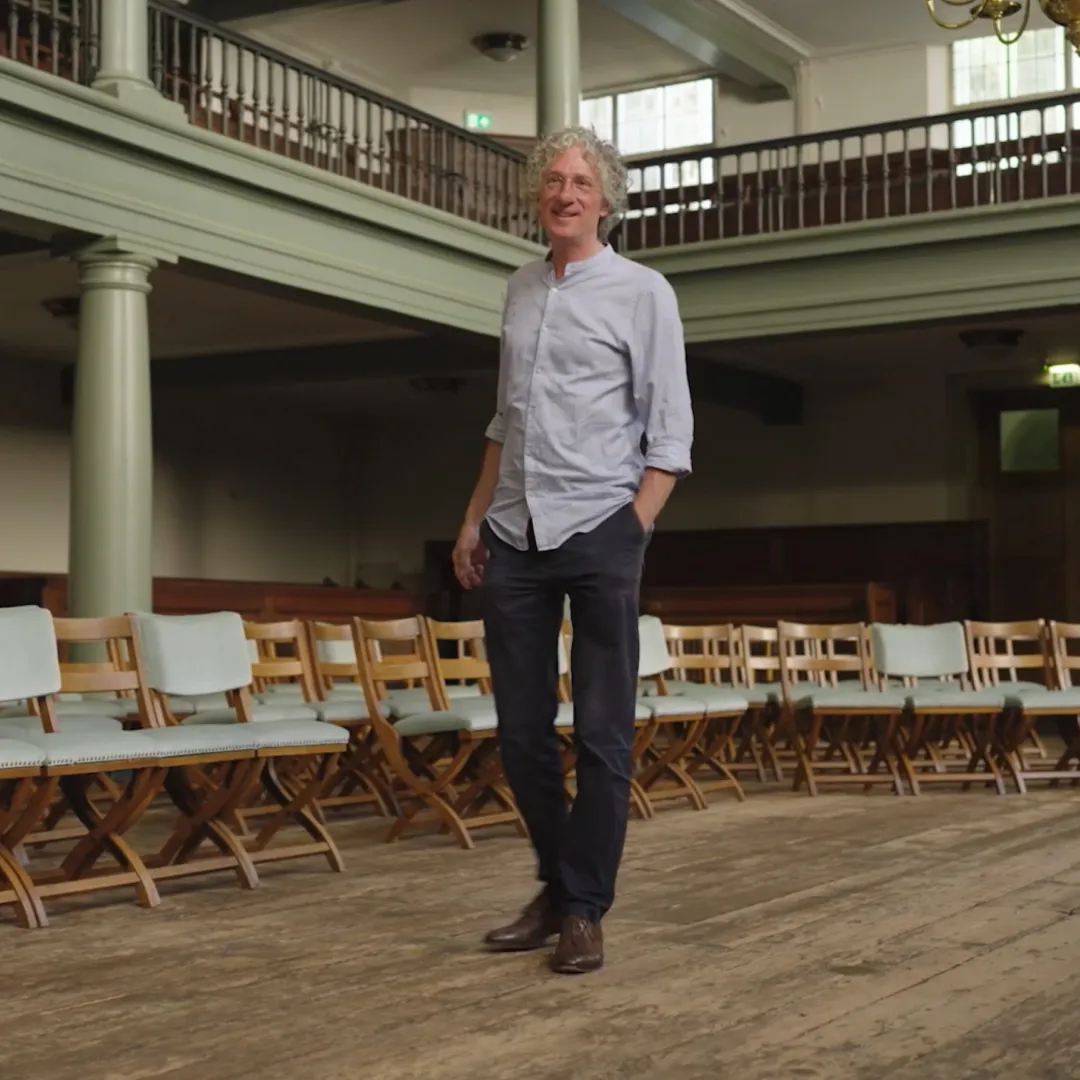 Man staat in een kerk zaal en kijkt naar de camera.