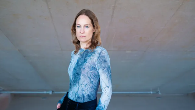 Laura Stek, een witte vrouw met lichtbruin haar, blauwe ogen en blauw shirt met lange mouwen, staat voor een betonnen muur en kijkt in de camera.