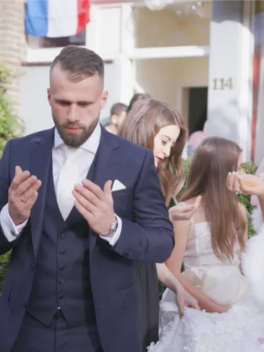 Een witte man in donkerblauw pak bidt met opgetilde handen voor een huis; naast hem staat een witte bruid in witte jurk en roze sluier, omringd door mensen, onder wie een zwarte vrouw in blauwe jurk.