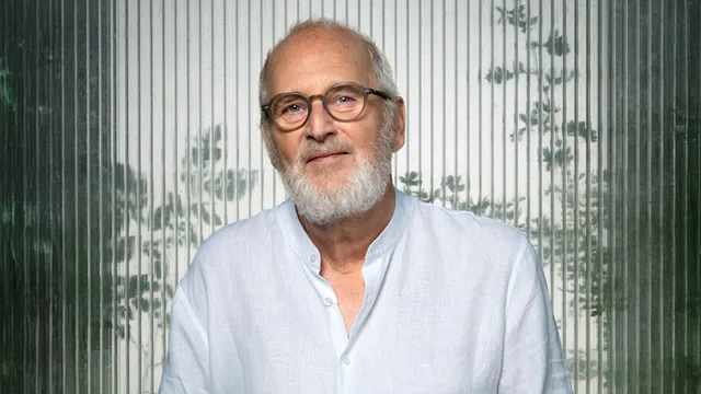 Een oudere man met een witte baard en bril draagt een witte linnen overhemd met opgerolde mouwen. Hij kijkt vriendelijk in de camera, staand voor een glazen wand waar vage silhouetten van takken en bladeren doorheen zichtbaar zijn.